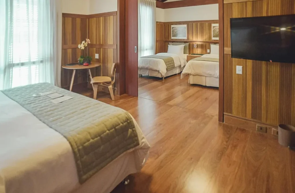 Acomodação familiar do Clara Ibiúna Resort com três camas, televisão, área de apoio e acabamento em madeira.
