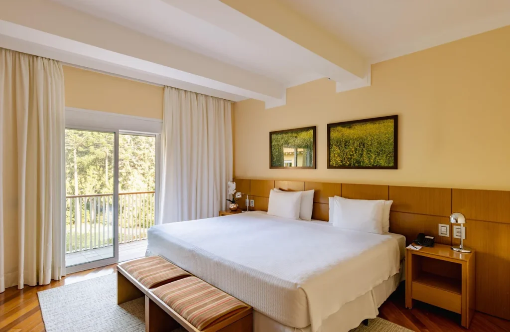 Quarto amplo e iluminado do Grande Hotel Campos do Jordão, com cama de casal, varanda e vista para a área verde.